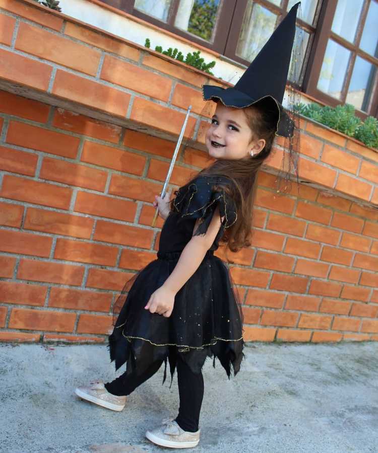 Garota fantasiada para o Halloween: Cores predominantes: preto. Usa: vestido, chapéu pontudo, meia calça, sapatilha, segura varinha mágica