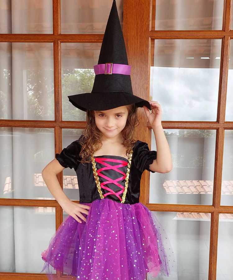 Garota com fantasia de bruxa infantil. Cores predominantes: preto e roxo. Usa: vestido e chapéu pontudo