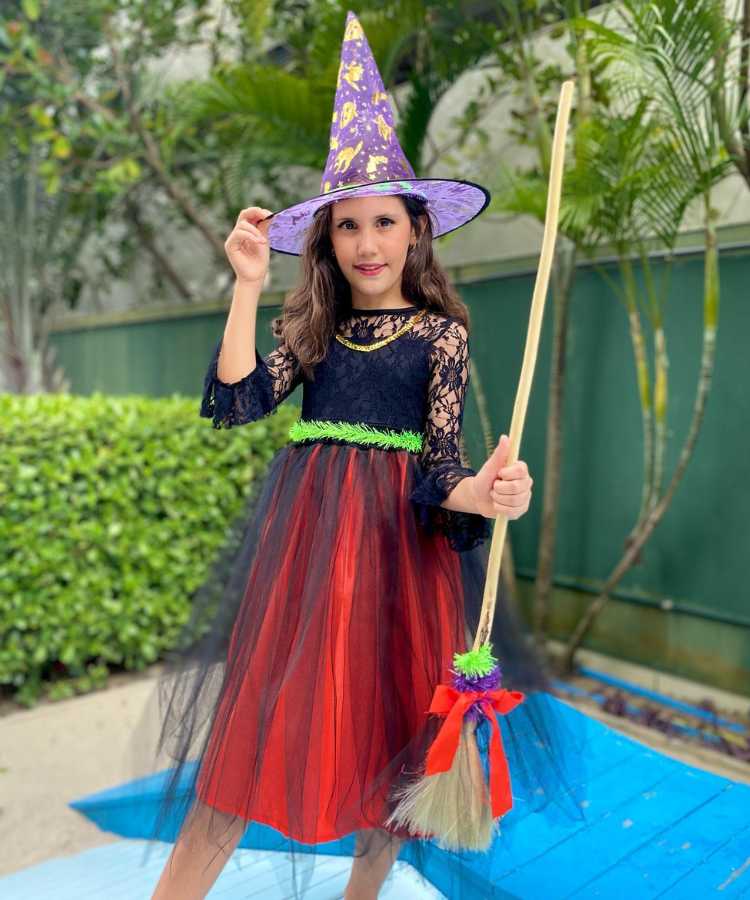 Garota fantasiada de bruxinha. Cores predominantes: vermelho, preto, lilás, verde. Usa: vestido até o joelho, chapéu transparente pontudo, segura vassoura de brinquedo
