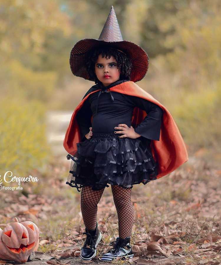 Garota fantasiada para o Halloween: Cores predominantes: preto e abóbora. Usa: blusa manga comprida, saia, meia arrastão, tênis, capa, chapéu pontudo