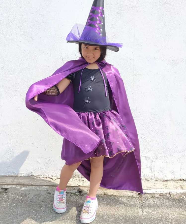 Garota fantasiada para o Halloween: Cores predominantes: preto e roxo. Usa: vestido, capa, chapéu pontudo, tênis