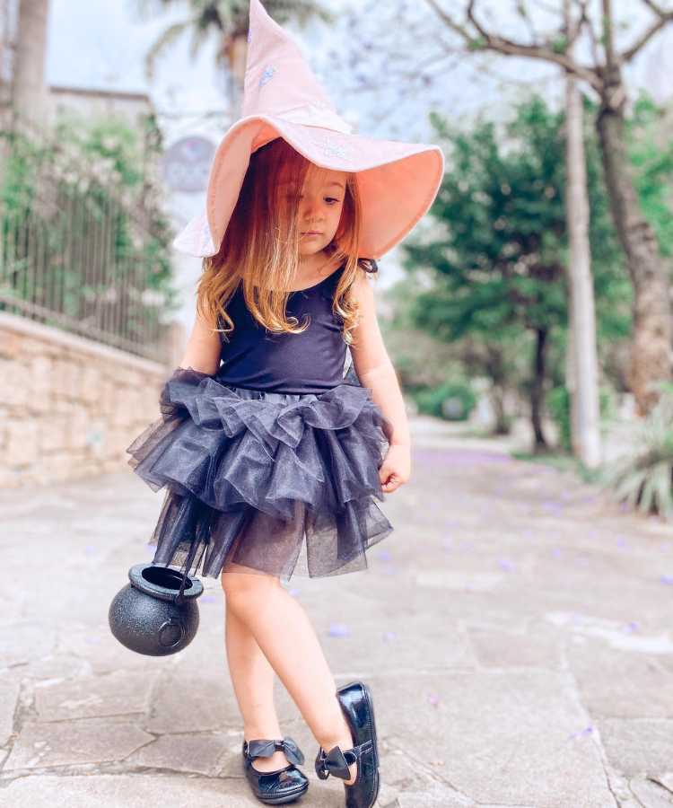 Garota fantasiada de bruxinha. Cores predominantes: preto e rosa. Usa: chapéu pontudo, vestido, sapatilha e baldinho em formato de caldeirão