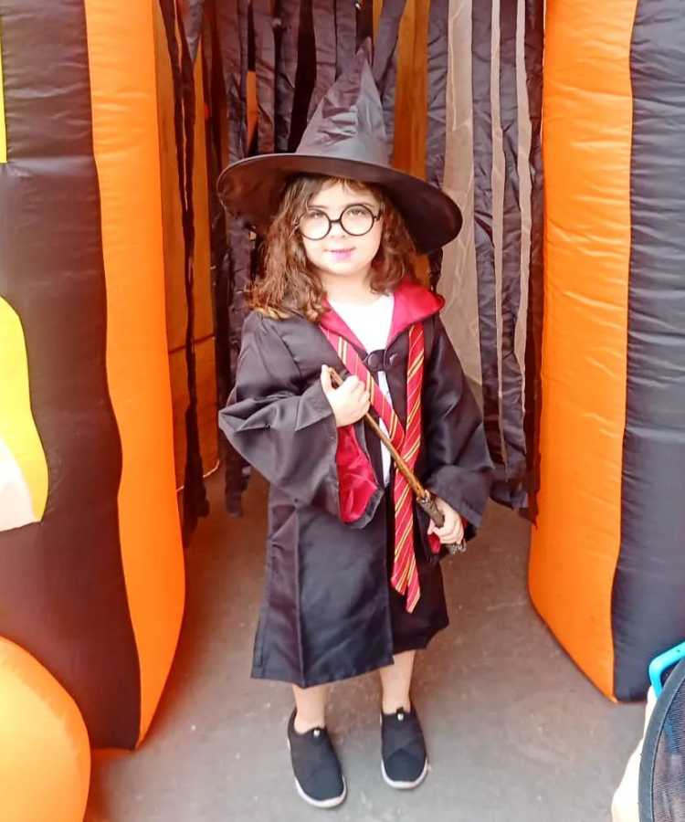 Garota fantasiada para o Halloween Harry Potter: Cores predominantes: vermelho, preto. Usa: capa com gravata, tênis, chapéu pontudo, segura varinha mágica
