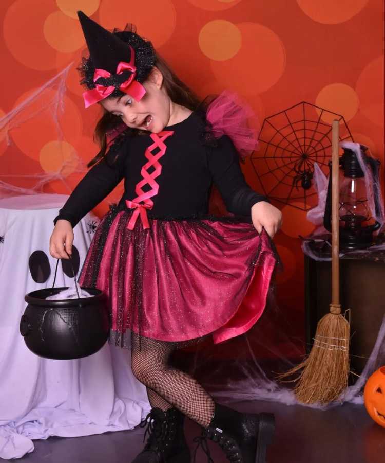 Garota com fantasia de bruxa infantil. Cores predominantes: prreto e pink. Usa: vestido manga comprida, meia calça arrastão, bota, tiara, mini chapéu pontudo + balde formato caldeirão