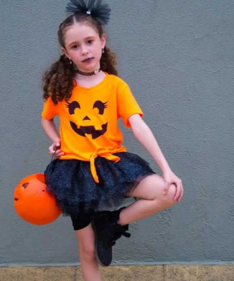 Garota fantasiada para o Halloween: Cores predominantes: preto e laranja. Usa: camiseta com cara de abóbora, saia de tule brilhante, tênis, tiara, balde em formato de abóbora