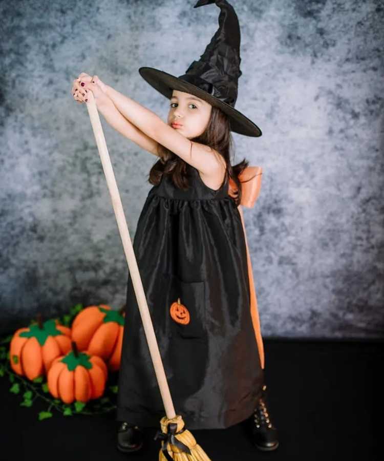 Garota fantasiada de bruxinha.. Cores predominantes: preto e abóbora. Usa: vestido longo com laço nas costas, chapéu pontudo, apoia-se em vassou de brinquedo