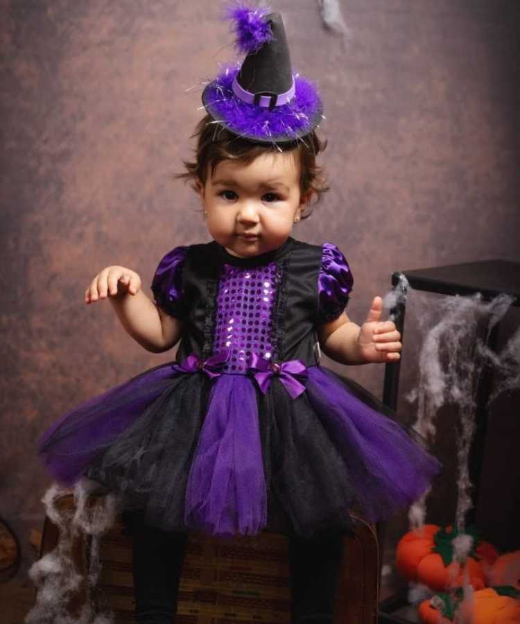 Bebê com fantasia de bruxa infantil. Cores predominantes: roxo e preto. Usa: vestido, meia calça, tiara mini chapéu pontudo