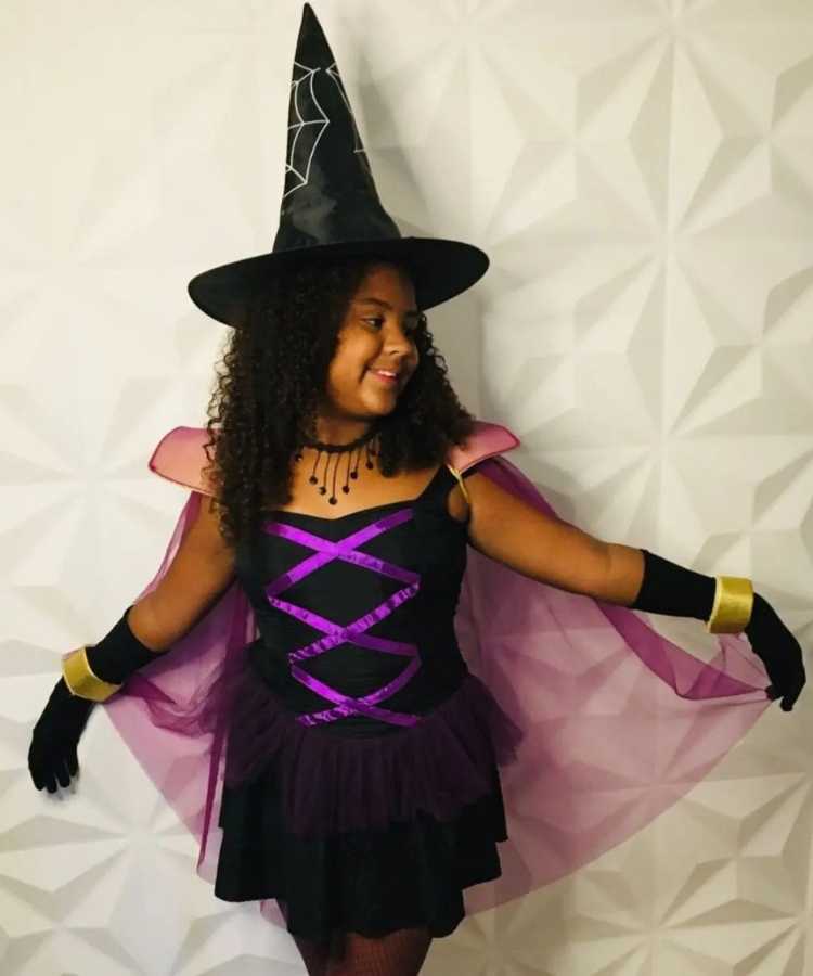 Garota fantasiada para o Halloween: Cores predominantes: preto, roxo. Usa: Chapéu pontudo,vestido com capa, luvas