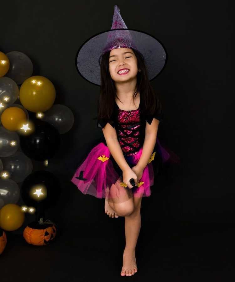 Garota fantasiada para o Halloween: Cores predominantes: preto e pink. Usa: chapéu pontudo, vestido e está com vassoura de brinquedo entre as pernas