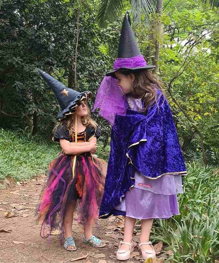 2 garotas com fantasia de bruxa infantil. Cores predominantes: roxo, preto, lilás, pink, abóbora. Usam: vestidos, chapéis pontudos, sandálias, uma tem capa