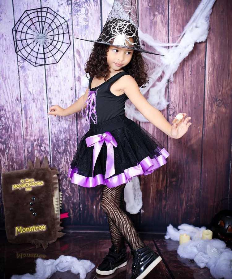Garota com fantasia de bruxa infantil. Cores predominantes: preto e roxo. Usa: chapéu pontudo, vestido, meia calça arrastão e tênis