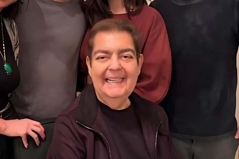 Foto de Faustão sentado usando roupa preta com outras pessoas atrás, que não aparecem