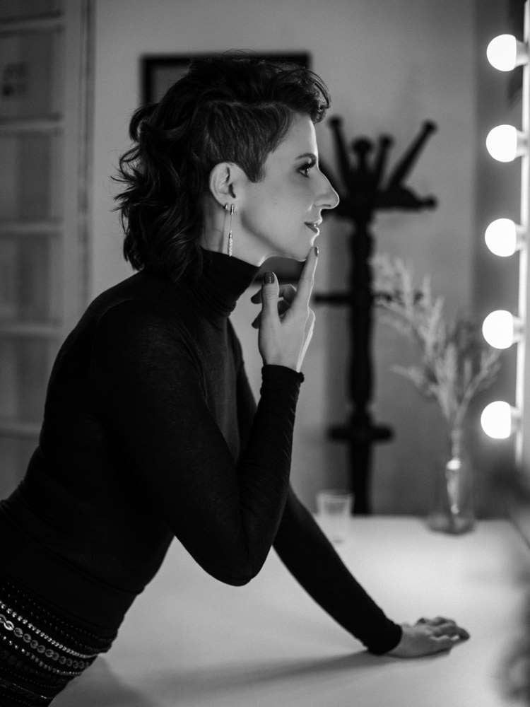 Mulher não-modelo com mais de 30 anos em ensaio fotográfico, usando body preto, brincos, se olhando no espelho de penteadeira. Foto preto e branco
