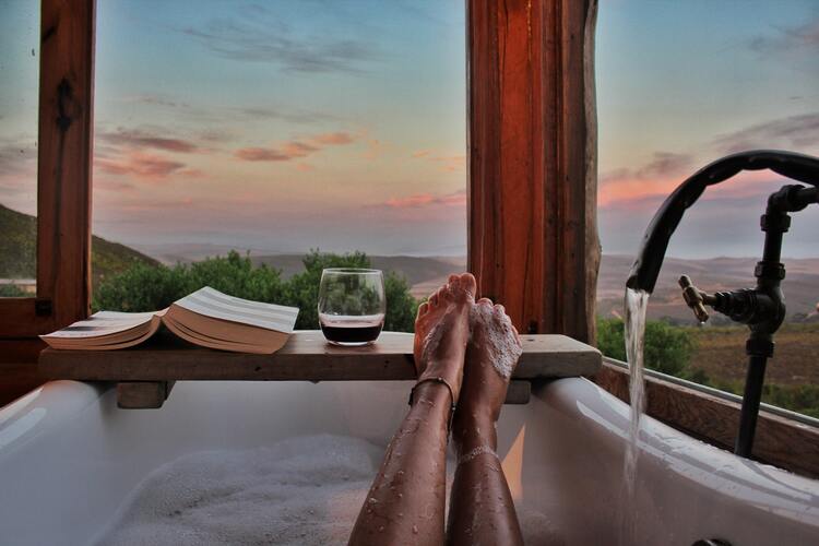 Mulher com pernas para cima em uma banheira de frente para uma janela com vista para natureza. Em cima das bordas da banheira há uma tábua de madeira que segura um livro e um copo com vinho.