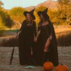 A imagem mostra duas pessoas vestidas de bruxas, com chapéus pontudos e capas pretas, em um campo ao ar livre. Elas estão cercadas por abóboras, sugerindo uma atmosfera de Halloween. Ao fundo, há árvores e montanhas sob a luz suave do entardecer, criando um cenário mágico e outonal.