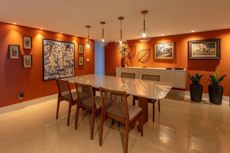 Sala de jantar com mesa grande, paredes em terracota, quadros e luminárias pendentes