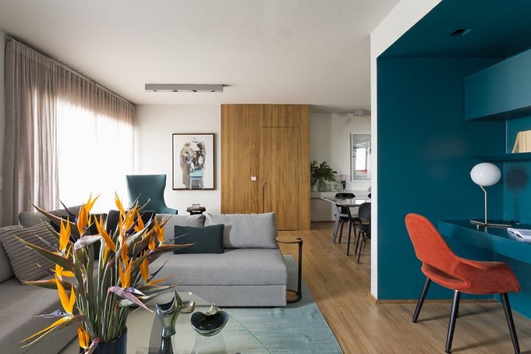 Sala de estar integrada com sala de jantar e home office. O piso e alguns detalhes são em madeira, sofá cinza, tapete verde e homme office em azul petróleo e cadeira laranja