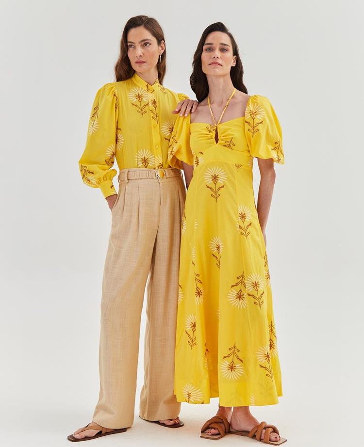 duas mulheres, a esquerda com calça solta bege e camisa de manga longa amarela, a direita com vestido longo amarelo de estampa florida