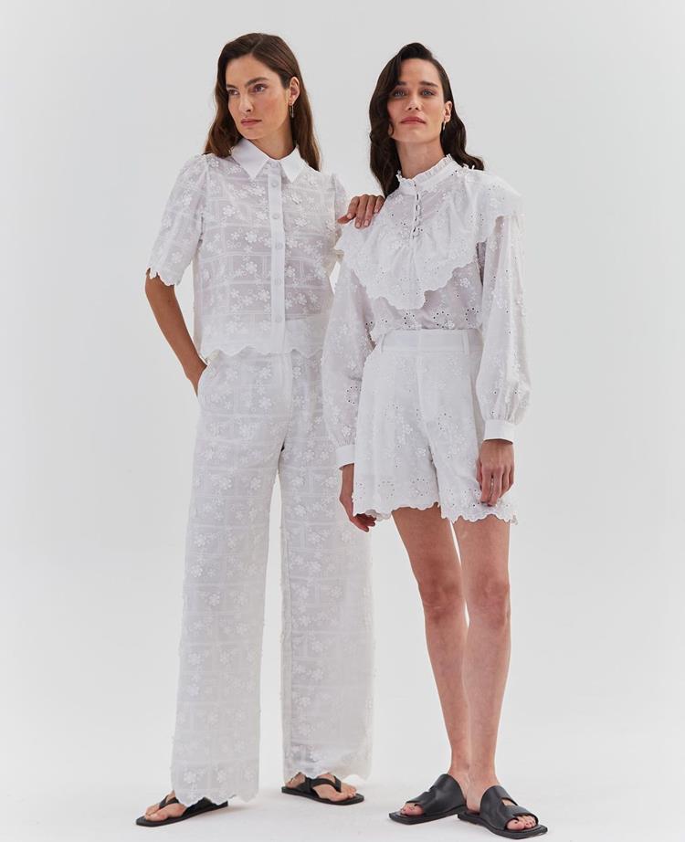 duas mulheres com conjunto branco rendado, sendo a da esquerda de calça e camisa de manga curta, e da direita de bermuda e camisa de manga comprida