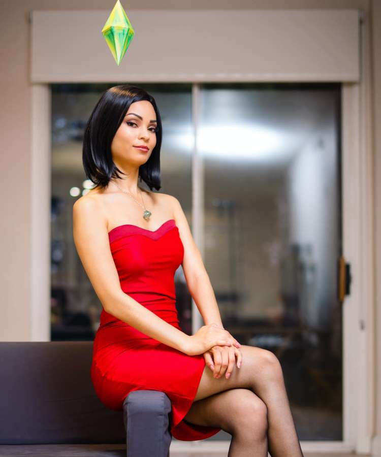 Fantasia, cosplay de mulher do The Sims, para o Halloween, usando tiara de prisma verde, vestindo vestido vermelho curto, sentada em sofá