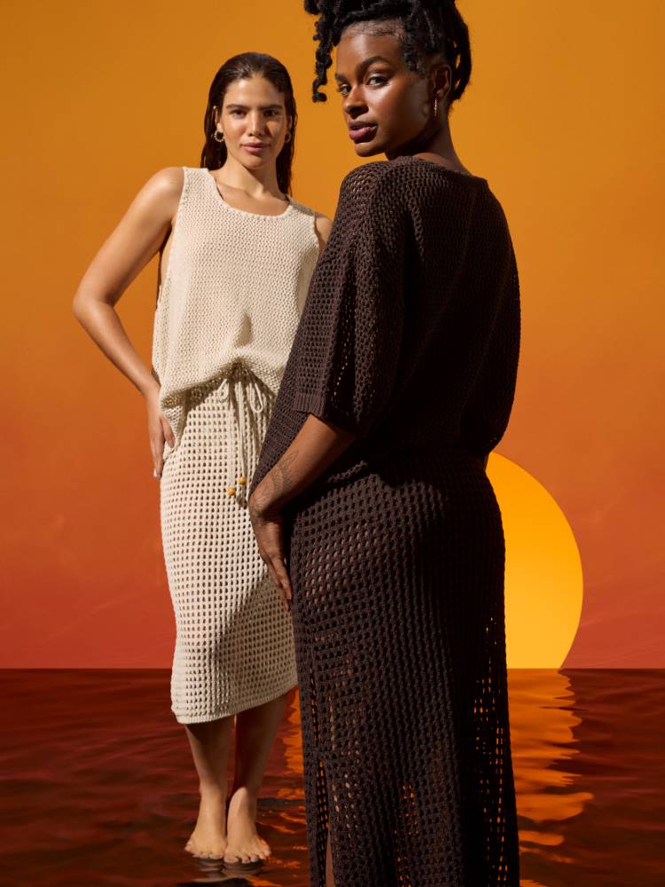 À esquerda, mulher de pele clara usando um conjunto de blusa de alça bege de crochê, assim como a saia mid. A mulher da direita possui pele negra e está de costas usando um conjunto de crochê marrom, de saia e blusa manga média. 