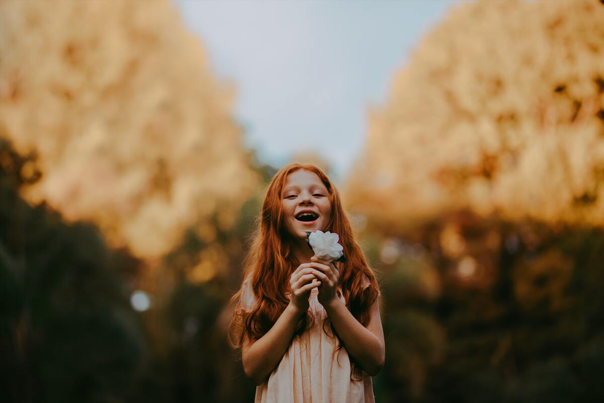 Criança ruiva do sexo feminino no meio de um jardim segurando uma flor branca na mão e sorrindo