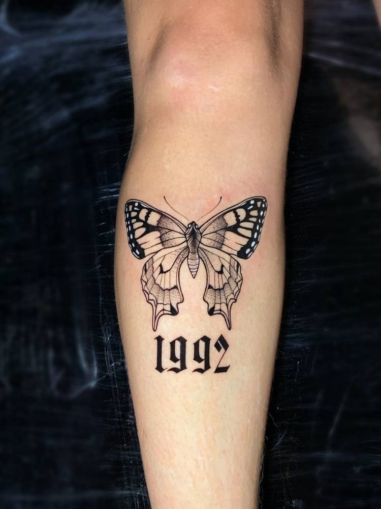 Tatuagem de borboleta na canela de uma pessoa de pele clara com sombreamento, traços finos e grossos e o número 1992 tatuado abaixo. 