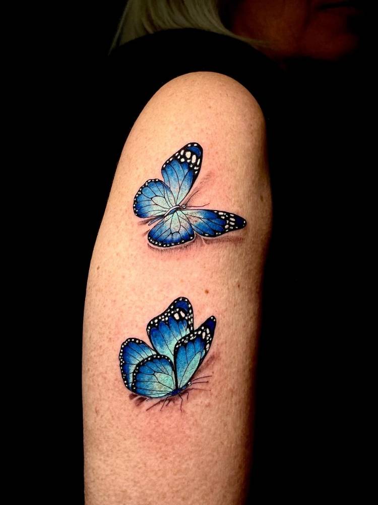 Braço de uma mulher de pele clara com duas borboletas delicadas azuis tatuadas com um leve sombreamento. 