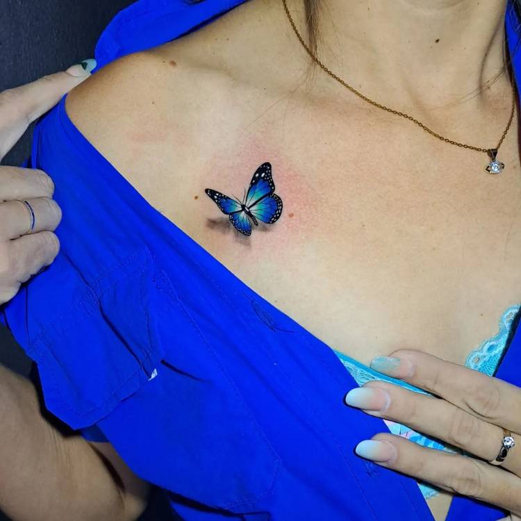 Pequena tatuagem de borboleta azul com sombreamento que dá efeito 3D de pouso do inseto no ombro de uma mulher de pele clara vestida com uma roupa azul. 