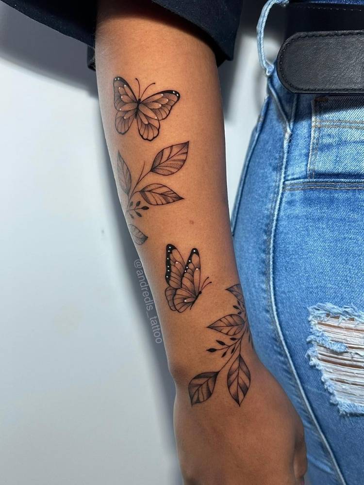 Tatuagem de borboleta com ramos em traço fino no braço de uma mulher com pele morena, usando blusa preta e calça jeans.