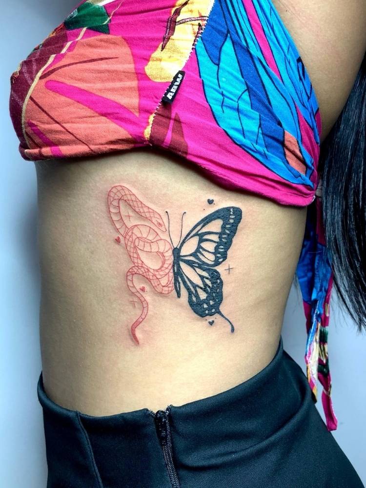 Tatuagem de uma asa de borboleta com traços grossos e pretos e a outra asa é o desenho de uma cobra de traços finos na cor vermelha. A tattoo foi feita na costela de uma mulher de pele clara, usando uma saia preta e uma blusa rosa, laranja e azul. 