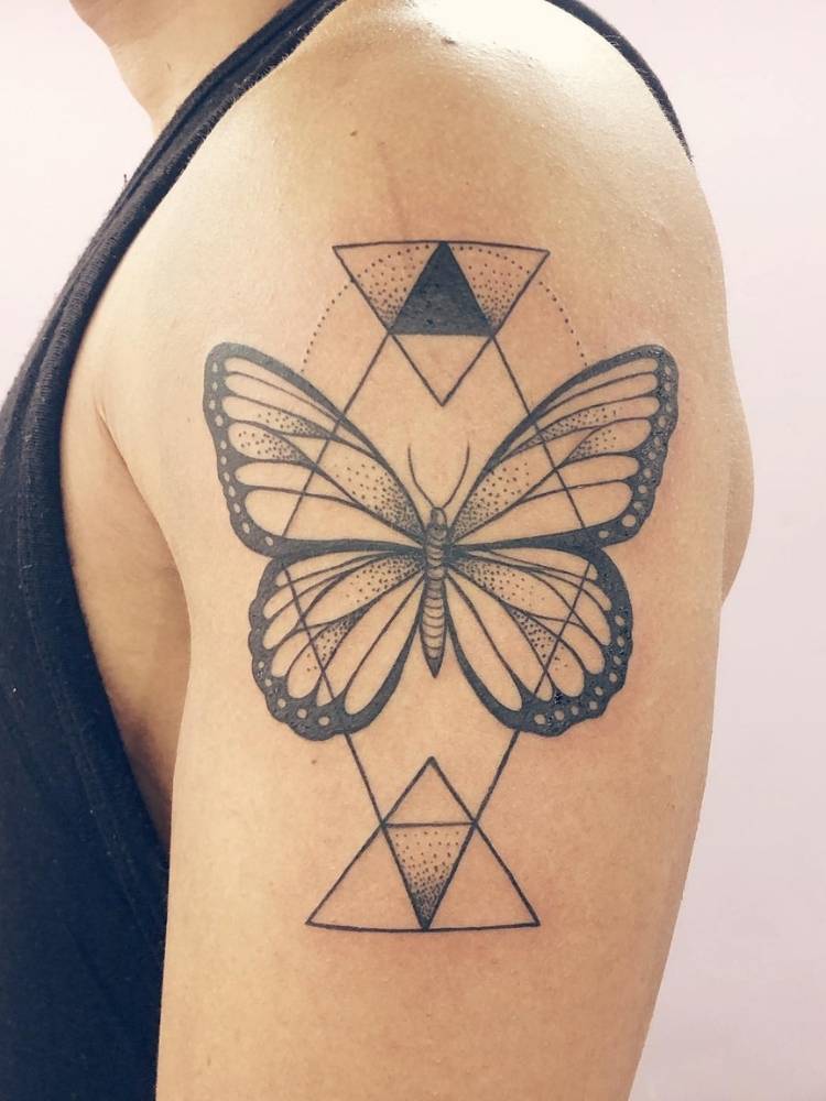 Tatuagem de borboleta com traços finos e grossos no  bíceps de homem de pele clara. Ela acompanha diversos traços triangulares.