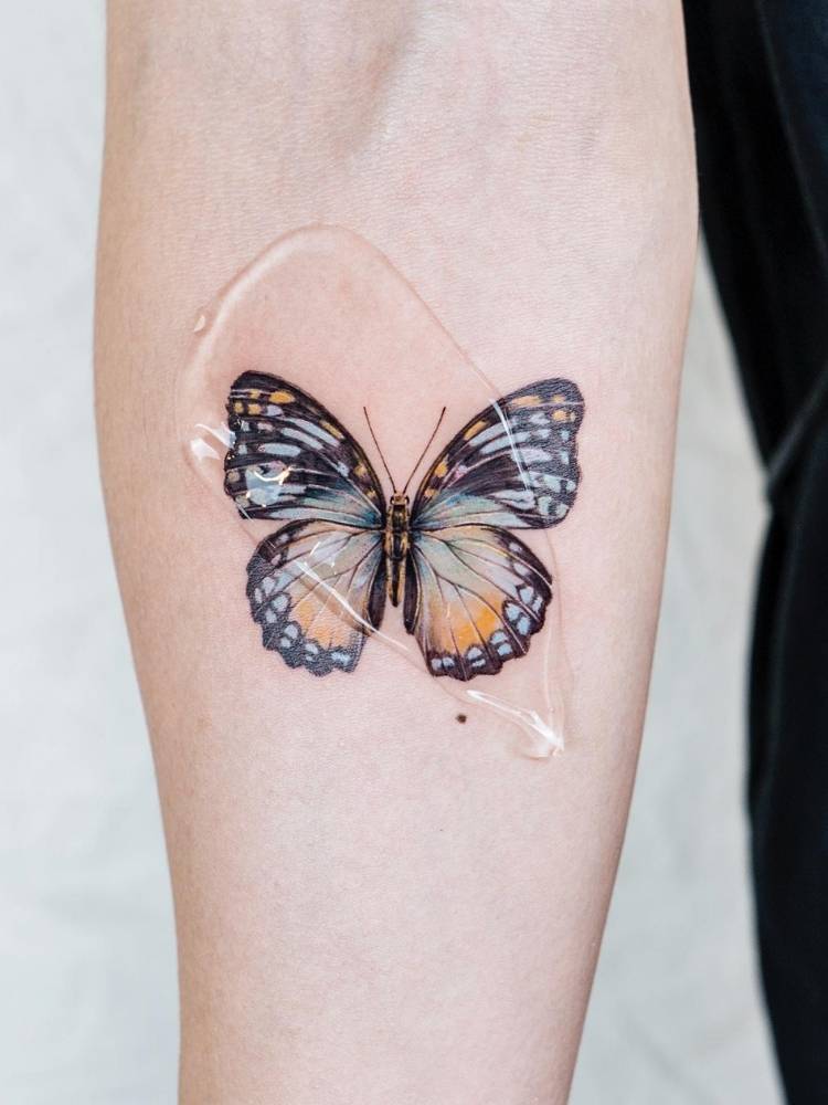 Tatuagem realista e cheia de detalhes de borboleta em tons de azul claro e amarelo no braço de uma pessoa de pele clara. 