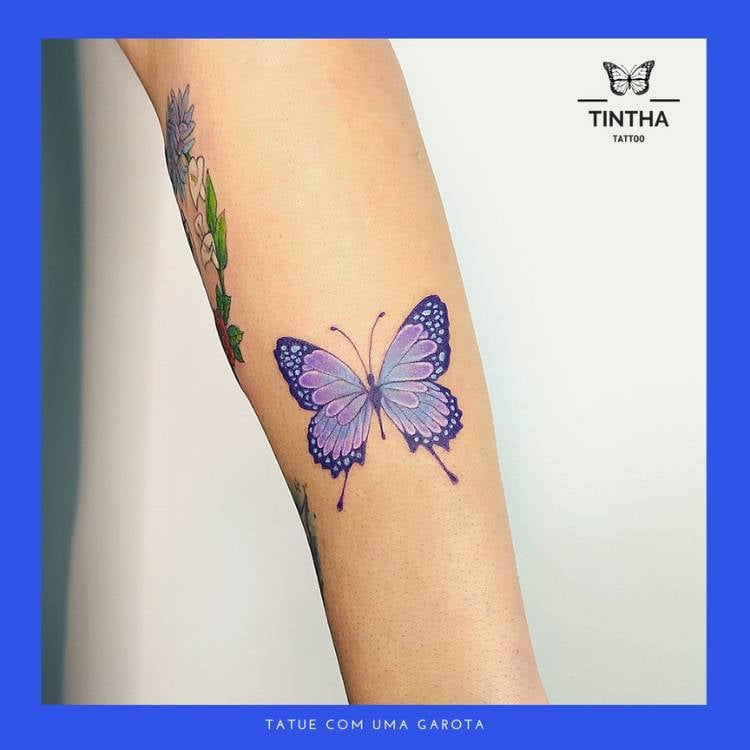 Tatuagem de borboleta em traços finos com tons de azul e roxo no braço de uma mulher de pele clara. 