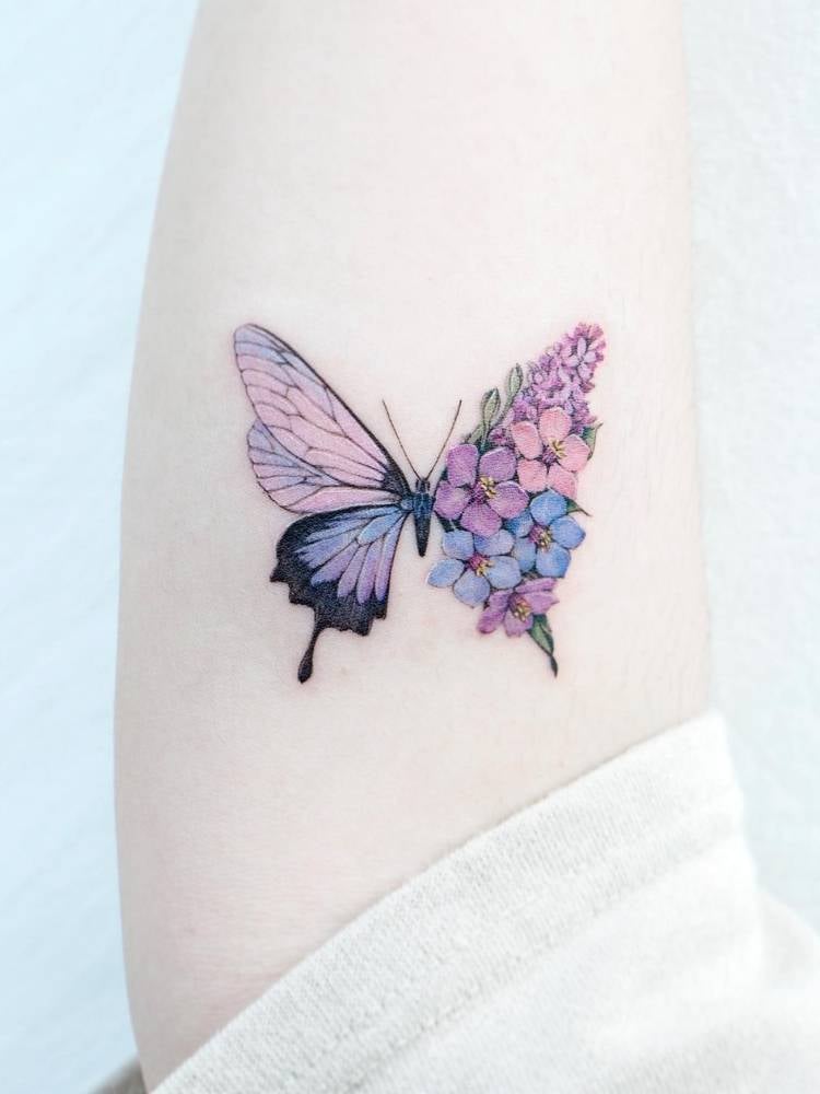 Tatuagem de borboleta azul, rosa e lilás cheia de detalhes. Feita no braço de uma mulher de pele clara. 