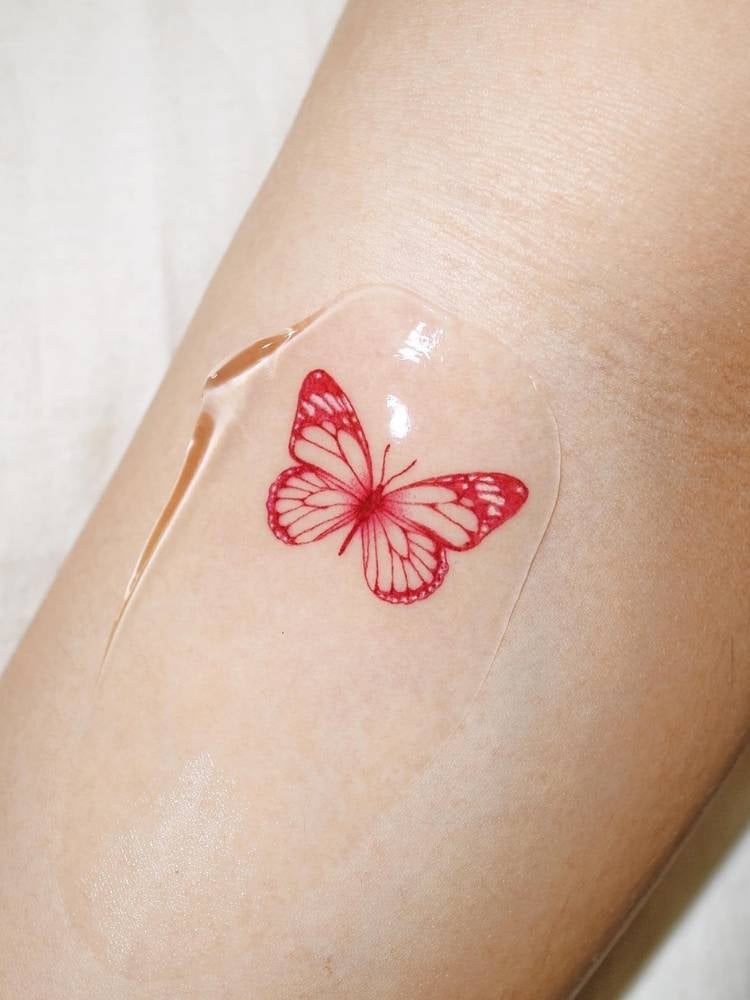 Tatuagem pequena de borboleta com traços finos e um pouco mais grossos em vermelho no braço de uma mulher de pele clara. 