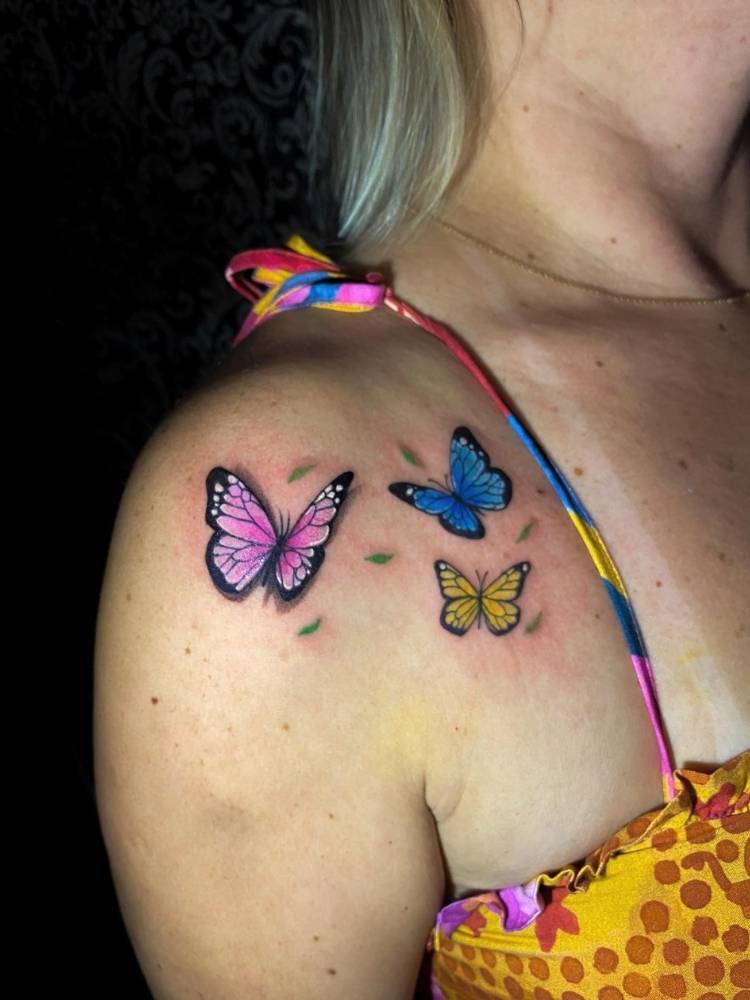 Mulher loira de pele clara com tatuagem de três borboletas coloridas no ombro, uma azul, outra roxa e outra amarela. 