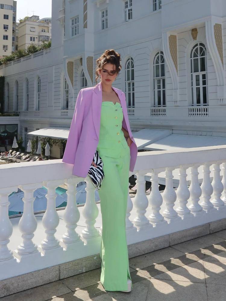 Mulher de pele clara em uma casa branca usando um conjunto de blusa e calça verde, sendo a peça se cima sem alça. Ela também usa um blazer lilás como sobreposição. 