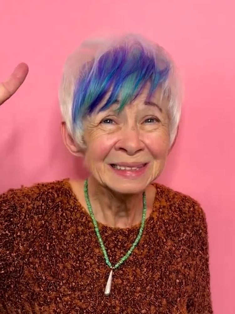 Mulher idosa com cabelo platinado e cor fantasia na franja, sorridente usando casaco marrom e colar verde de pedrinhas
