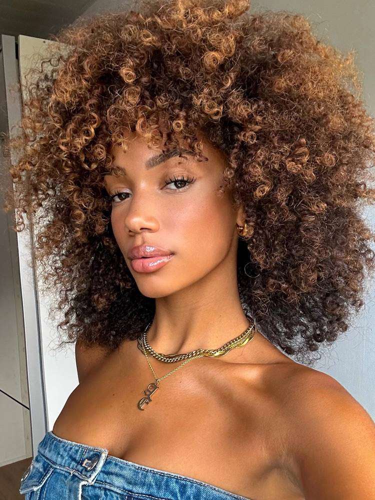Mulher com corte de cabelo afro fofo, uma das tendências do verão 2024. Ela tem o tom de pele escuro, usa tomara que caia jeans, joias douradas e tem o cabelo castanho iluminado