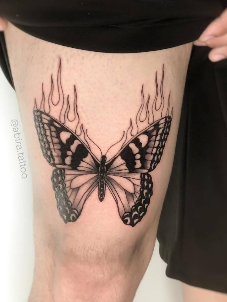 Tatuagem grande de borboleta na coxa de pele clara com sombreamento, traços grossos e detalhes de chamas. 