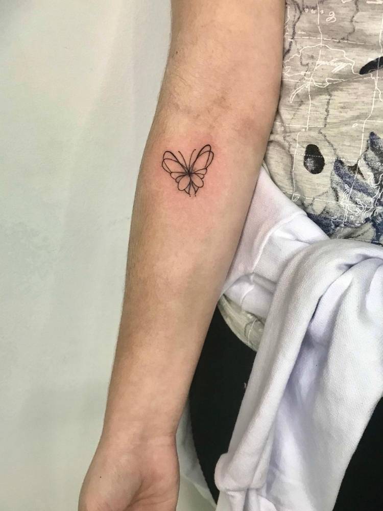 Tatuagem de borboleta delicada com poucos traços, sendo eles finos e pretos, pequena e minimalista. Feita no braço de uma mulher de pele clara. 
