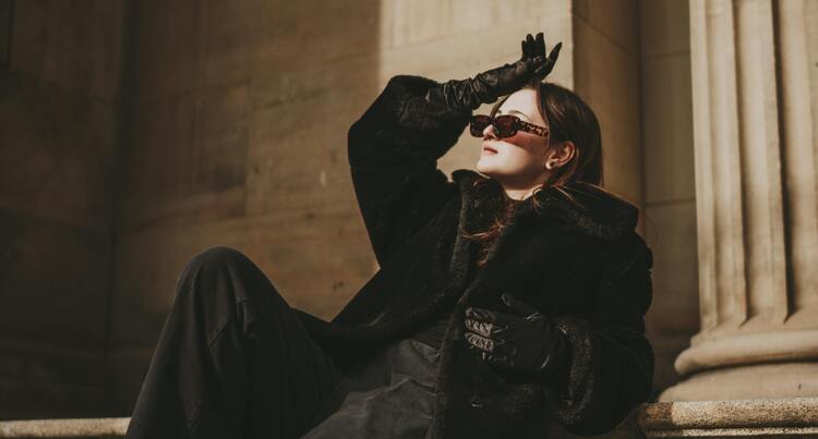 Mulher com roupas pretas e luvas sentada em uma escadaria usando óculos de sol e olhando para cima