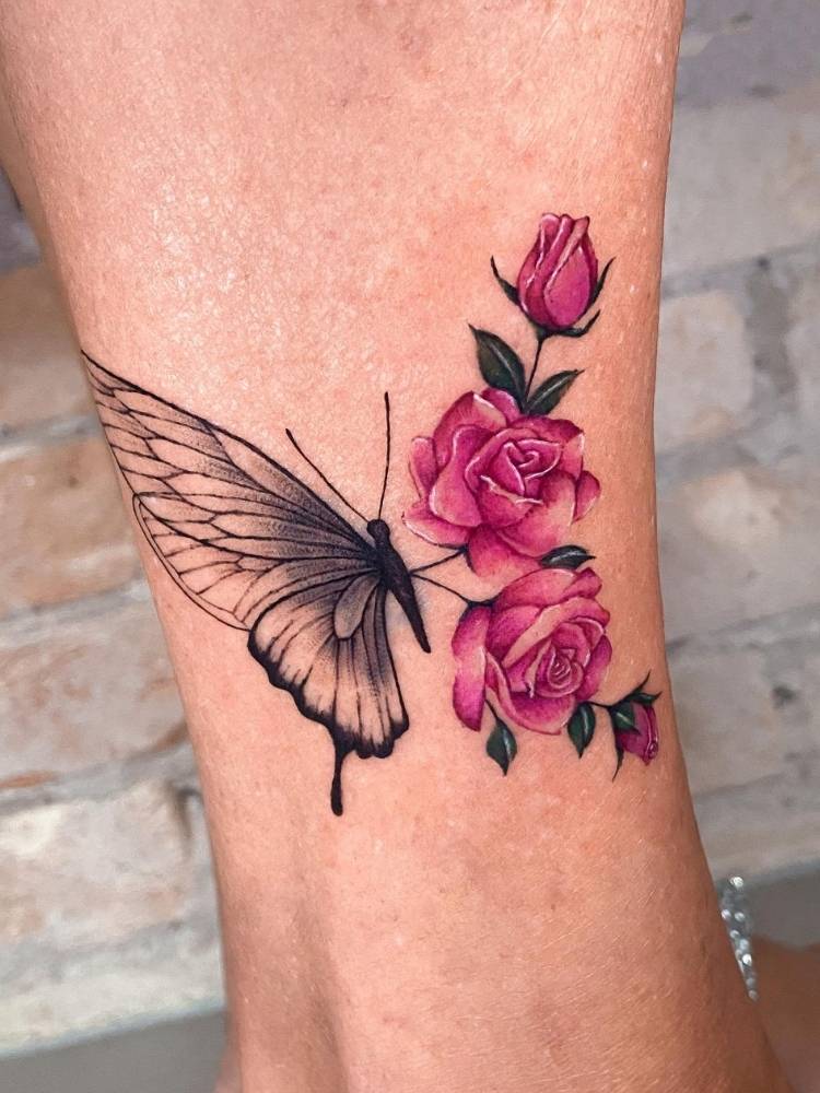 Tatuagem de borboleta com asa metade em traços finos pretos com sombreado e a outra metade com flores rosas. Feita no braço de uma mulher de pele clara. 