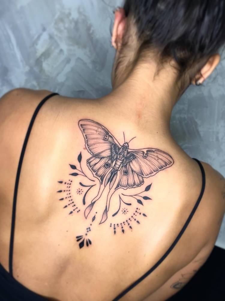 Tatuagem grande e chamativa de uma borboleta com traços grossos, sombreada, ramos e arabescos. nas costas de uma mulher de pele morena e cabelo preso. 