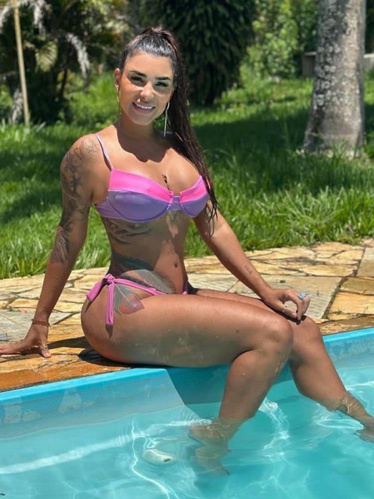 Jenny Miranda de biquíni roxo e rosa sentada na beirada da piscina.