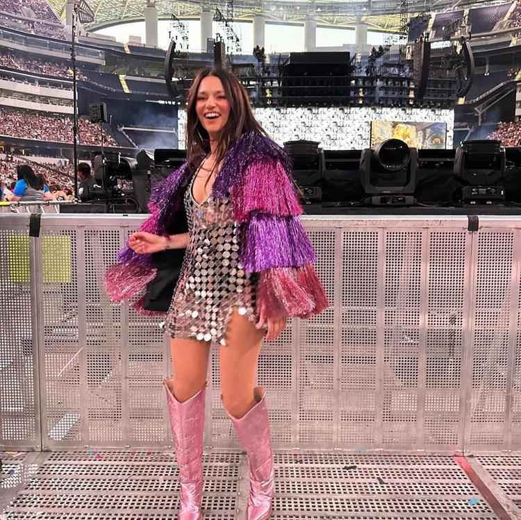 mulher em estádio com vestido curto brilhoso prata, casaco de franjas colorido em tons de rosa com brilhos e bota longa rosa