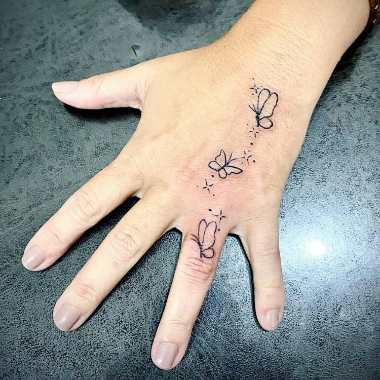 Tatuagem de várias borboletas pequenas no dorso e dedo anelar da mão de uma mulher de pele clara. 