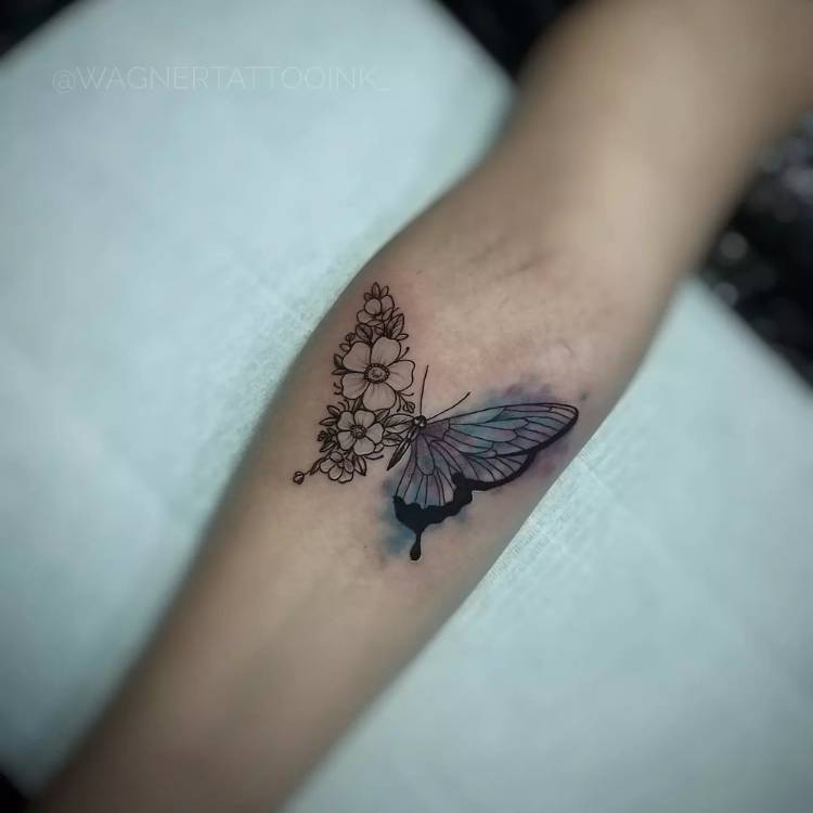 Tatuagem de borboleta no braço de uma mulher de pele clara sendo metade da asa em flores de traço fino e a outra metade uma asa em estilo aquarela em azul. 