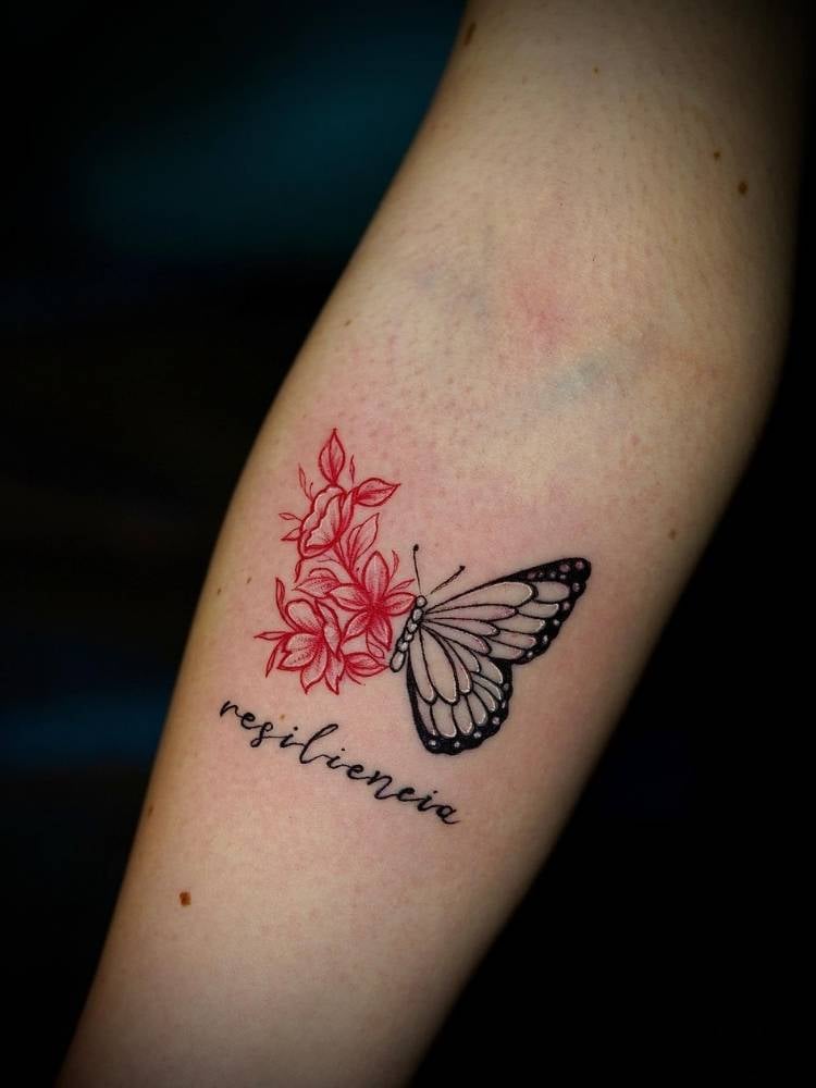 Tatuagem de borboleta no braço de uma mulher de pele clara. Metade da asa do inseto são flores em traço fino vermelho e a outra metade é uma asa normal com traços finos e grossos. Abaixo do desenho, há a palavra resiliência.